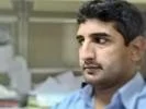 A man with short dark hair and a mustache, wearing a light blue shirt, sitting in an office environment with blurred papers and shelves in the background