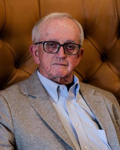 An elderly man with short white hair, wearing glasses, a light blue shirt, and a beige blazer, sitting against a tufted leather backdrop.