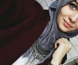 A close-up of a woman wearing a gray hijab and a maroon jacket, accessorized with an intricate silver necklace. She is smiling softly with a relaxed and elegant pose