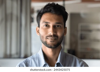 A portrait of a man with a trimmed beard and short hair, wearing a light blue shirt, standing in a modern office environment with a neutral expression.
